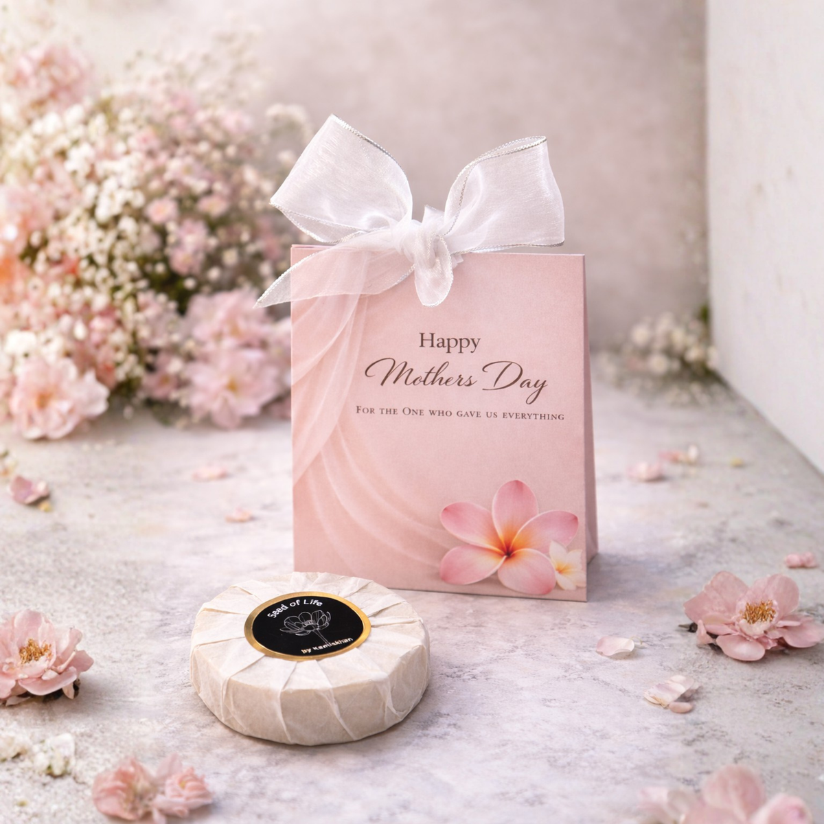 Pink 'Happy Mother's Day' gift box soap with a white bow, surrounded by flowers on a light surface.