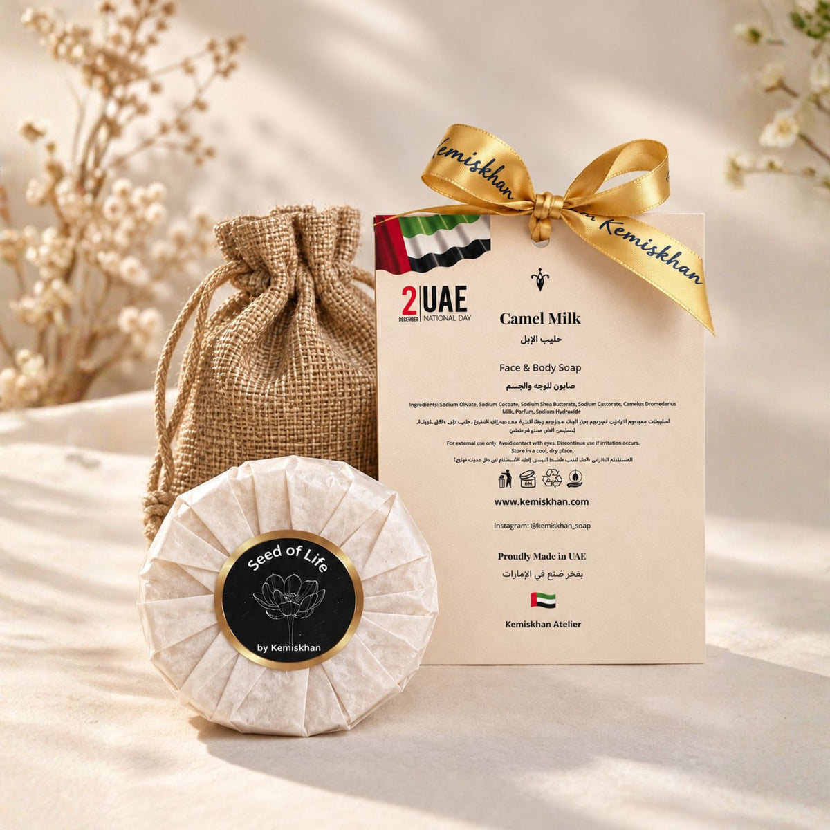 Camel milk face and body soap by Seed of Life by Kemishkhan, shown wrapped in textured paper with a circular black label, next to its burlap drawstring pouch and branded card tied with a gold ribbon, on a beige fabric background.