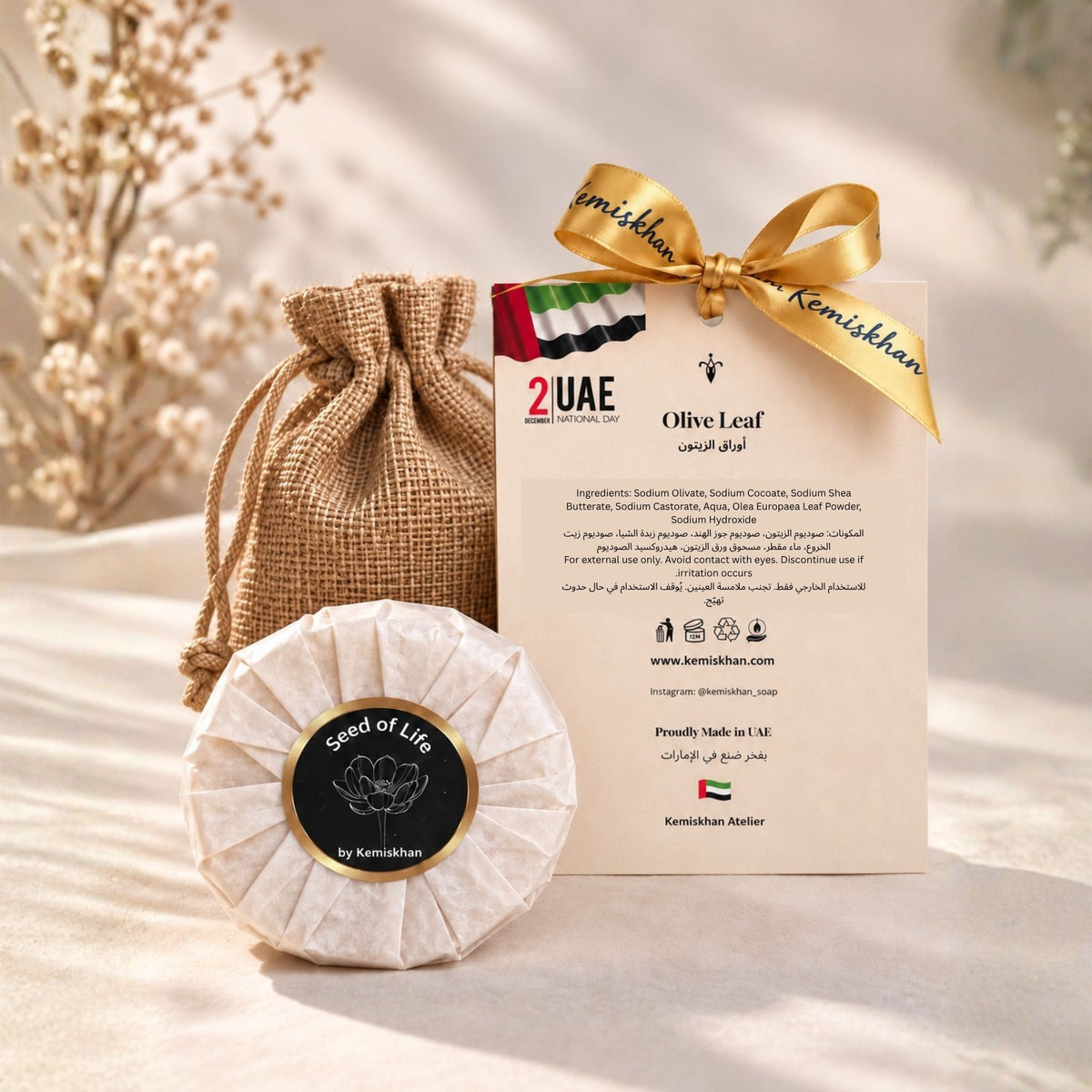 Olive Leaf face and body soap by Seed of Life by Kemishkhan, shown wrapped in textured paper with a circular black label, next to its burlap drawstring pouch and branded card tied with a gold ribbon, on a beige fabric background.