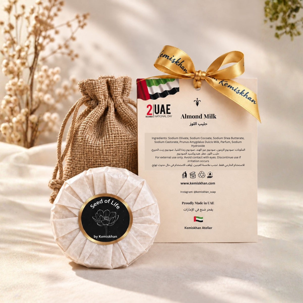 Almond Milk Gift face and body soap by Seed of Life by Kemishkhan, shown wrapped in textured paper with a circular black label, next to its burlap drawstring pouch and branded card tied with a gold ribbon, on a beige fabric background.