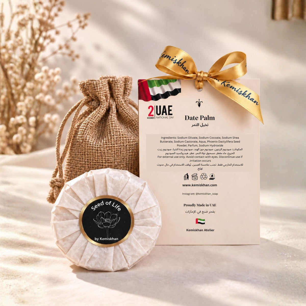 Date Palm face and body soap by Seed of Life by Kemishkhan, shown wrapped in textured paper with a circular black label, next to its burlap drawstring pouch and branded card tied with a gold ribbon, on a beige fabric background.